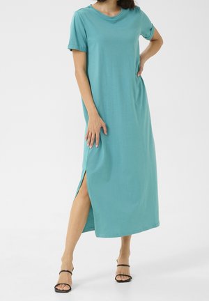 Vrouw met een teal midi-jurk met korte mouwen en een zijsplit, poserend met één hand op haar heup en zwarte sandaaltjes met bandjes.