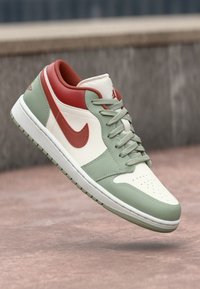 Baskets basses avec des panneaux blancs, vert sauge et rouge profond, un swoosh Nike rouge et des lacets assortis sur une surface en béton.