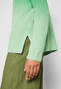 Gros plan d'une personne portant un pull tricoté vert clair avec des fentes latérales et une jupe vert olive, montrant sa main droite détendue le long du corps.