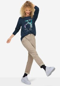 Maglietta a maniche lunghe blu con grafica animale, abbinata a pantaloni chino beige chiaro e sneakers bianche. Capelli ricci, mani alzate, posa dinamica.