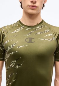 Νεαρό άτομο που φοράει στενή πουκαμίσα ελιάς με αφηρημένες ανοιχτόχρωμες ρίγες και το κεντρικό λογότυπο Champion στο στήθος.