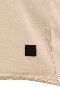Beige Stoff mit glatter Textur, versehen mit einem schwarzen Quadratlabel mit erhabenem Text, das sich nahe der Unterseite befindet.