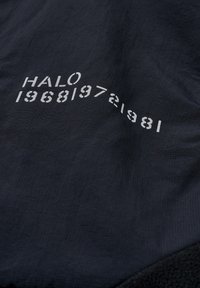 Svart textil med tryckt vit text: "HALO 196819721981." Slät yta med subtila horisontella linjer; kontrasterar med en mjuk svart tygkant.