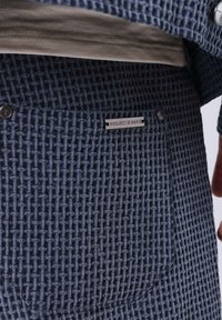 Pantalon bleu texturé avec un motif tissé, portant une étiquette affichant "PROJECT X PARIS" et deux boutons décoratifs sur la ceinture.