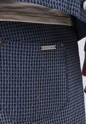 Pantaloni blu con motivo intrecciato e trama testurizzata, con un'etichetta che recita "PROJECT X PARIS" e due bottoni decorativi sulla vita.