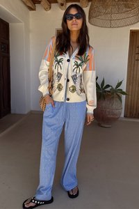 White cardigan with colorful palm embroidery and orange stripes, paired with blue striped pants, worn with black sandals. Natural background.