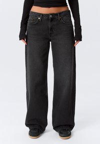 Personne portant un jean noir ample et des baskets noires, debout avec les mains près des hanches sur un fond clair uni.