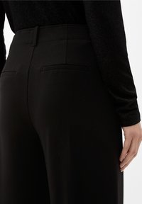 Black high-waisted trousers with a smooth texture, featuring two back pockets and a tailored fit for sleek lines.