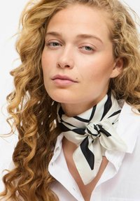 Écharpe en soie avec un motif noué en cr�ème et rayures noires. Le modèle la porte autour du cou, assortie à une chemise blanche.