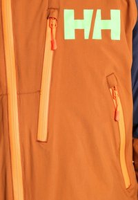 Veste marron à fermeture éclair avec accents orange et vert, dotée de deux poches zippées et d'un grand logo "HH". Le matériau semble léger et lisse.