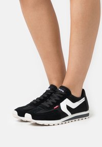 Ben som bär svarta och vita Levi's sneakers med snörning, står på en vit yta.