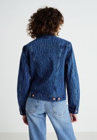 Chaqueta de mezclilla azul oscura con patrón ondulado en relieve, detalles de botones y un cuello clásico. El corte es ligeramente holgado, con bolsillos en la parte trasera.