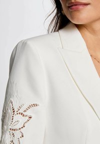 Blazer bianco con ricami intricati sulle maniche. Presenta una texture liscia e rever strutturati. Incentrato su una vestibilità sartoriale.