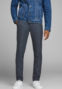 Pantaloni slim-fit grigio scuro, con una texture e una finitura liscia. Indossati con una giacca in denim blu e sneakers bianche.