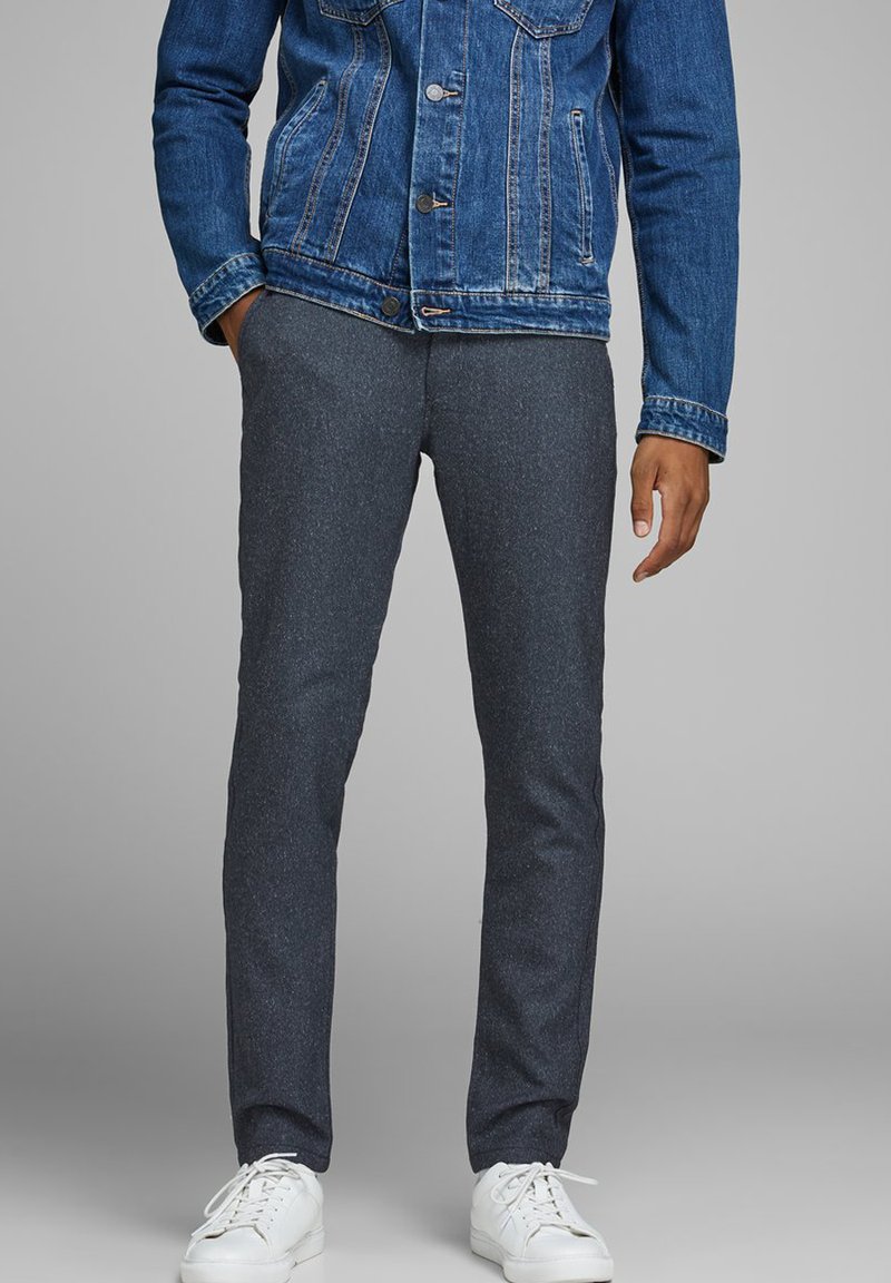 Pantaloni slim-fit grigio scuro, con una texture e una finitura liscia. Indossati con una giacca in denim blu e sneakers bianche.