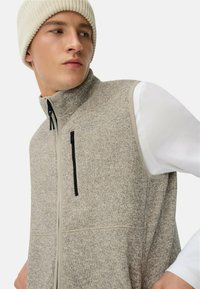 Graues Fleecewesten mit hohem Kragen und Frontreißverschluss. Verfügt über eine Reißverschlusstasche und schwarze Akzente, übereinander getragen über einem weißen Langarmshirt.