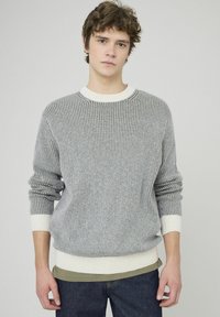 Pull en maille côtelée gris avec des poignets et un ourlet blancs, avec un col rond et des manches longues, porté par-dessus une chemise verte.