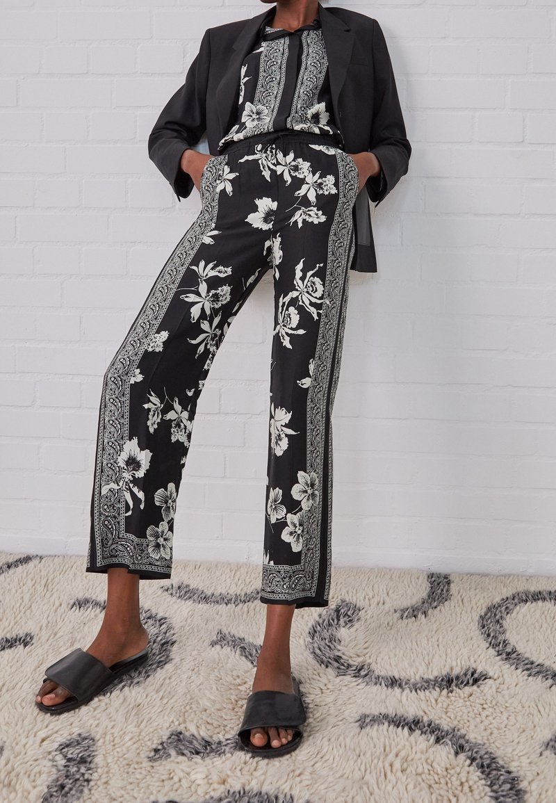 Personne portant un pantalon à motifs floraux noirs et un haut assorti, avec un blazer noir et des sandales noires, debout sur un tapis texturé.