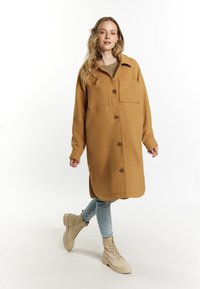 Cappotto oversize color beige realizzato in tessuto morbido, con colletto, due tasche frontali e bottoni marroni. Abbinato a jeans azzurri chiari e stivali beige.