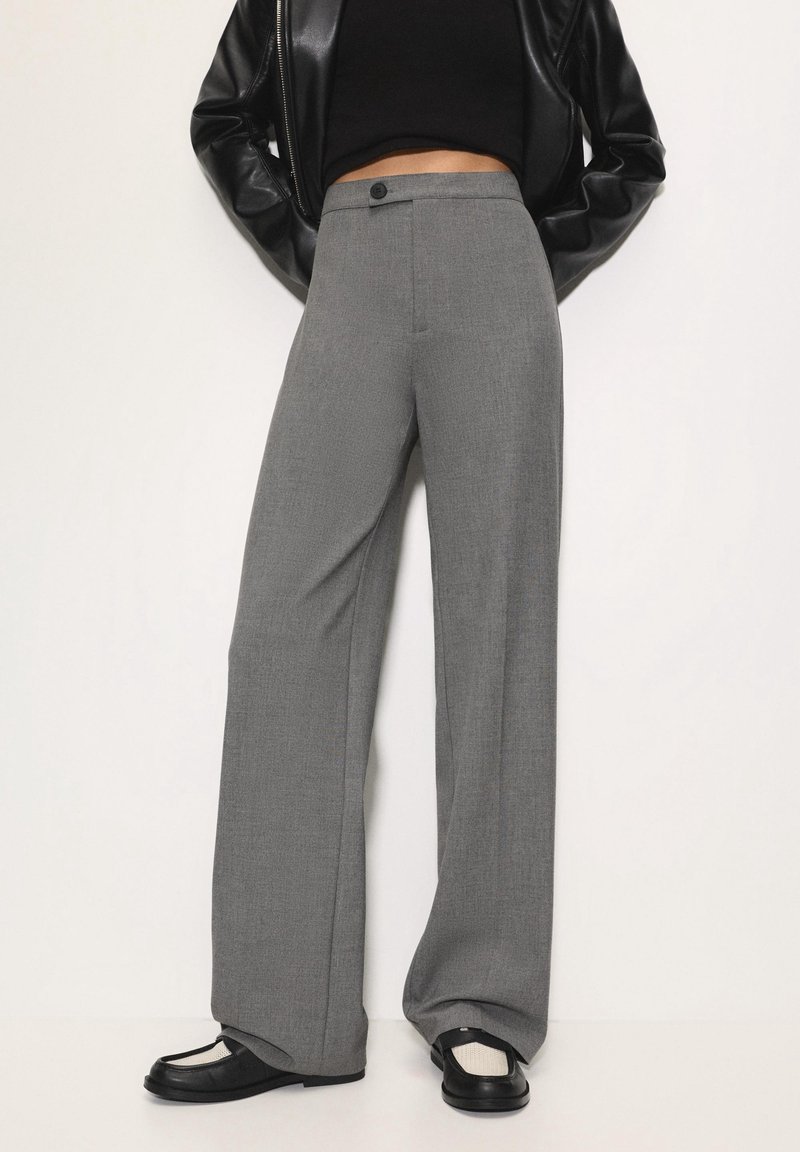 Personne portant un pantalon gris taille haute à jambes larges, un top court noir, une veste en cuir noire et des chaussures noires avec des chaussettes blanches sur un fond uni.