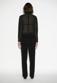 Schwarzes, durchscheinendes Oberteil aus strukturiertem Stoff mit gepunktetem Muster, cropped Design, kombiniert mit schwarzen Hosen, vor einem schlichten weißen Hintergrund stehend.