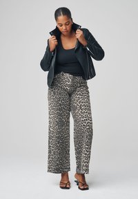 Veste en cuir noir, débardeur noir et pantalon large à imprimé léopard avec un fond beige clair. Sandales noires à brides.