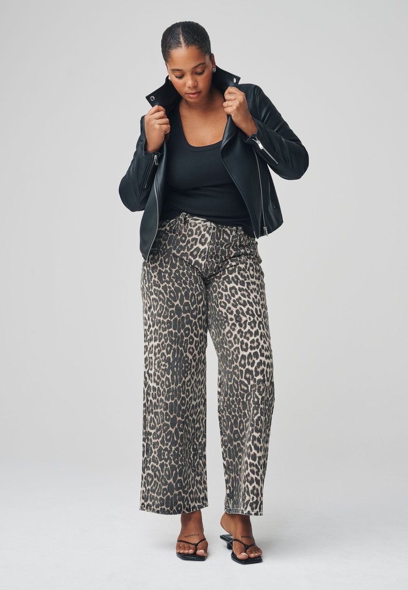 Veste en cuir noir, débardeur noir et pantalon large à imprimé léopard avec un fond beige clair. Sandales noires à brides.