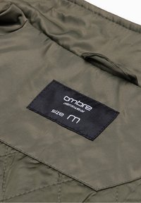 Étiquette de vêtement noire sur tissu matelassé olive, indiquant « ombre menswear taille M » avec une boucle en tissu au-dessus de l'étiquette.