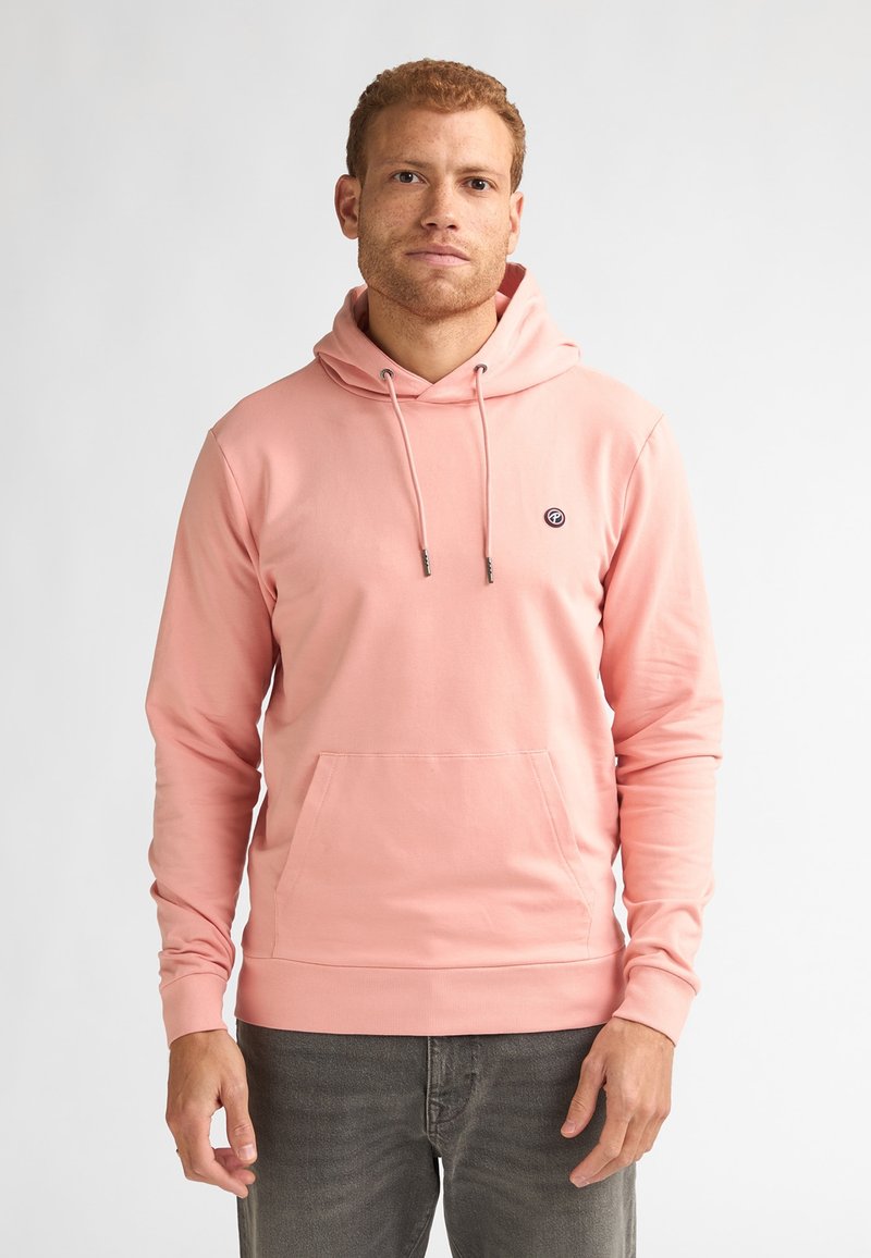 Homme portant un sweat à capuche rose avec poche avant et cordons de serrage, associé à un jean gris foncé, debout devant un fond blanc.