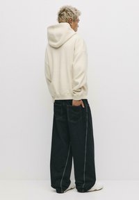 Felpa con cappuccio bianca a maniche lunghe e vestibilità ampia, abbinata a jeans neri larghi con cuciture bianche. Modello mostrato di spalle, indossa sneakers bianche.