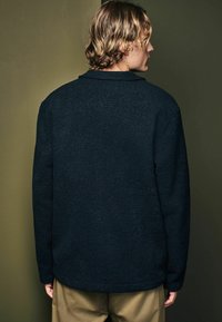 Dunkelblaue Fleece-Jacke mit Kragen, mit strukturiertem Oberflächenmaterial. Getragen mit khakifarbenen Hosen, Ansicht von hinten.