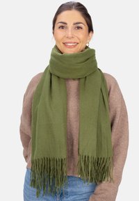 Écharpe vert olive en tissu doux, avec des bords frangés. Portée lâchement autour du cou sur un pull marron.