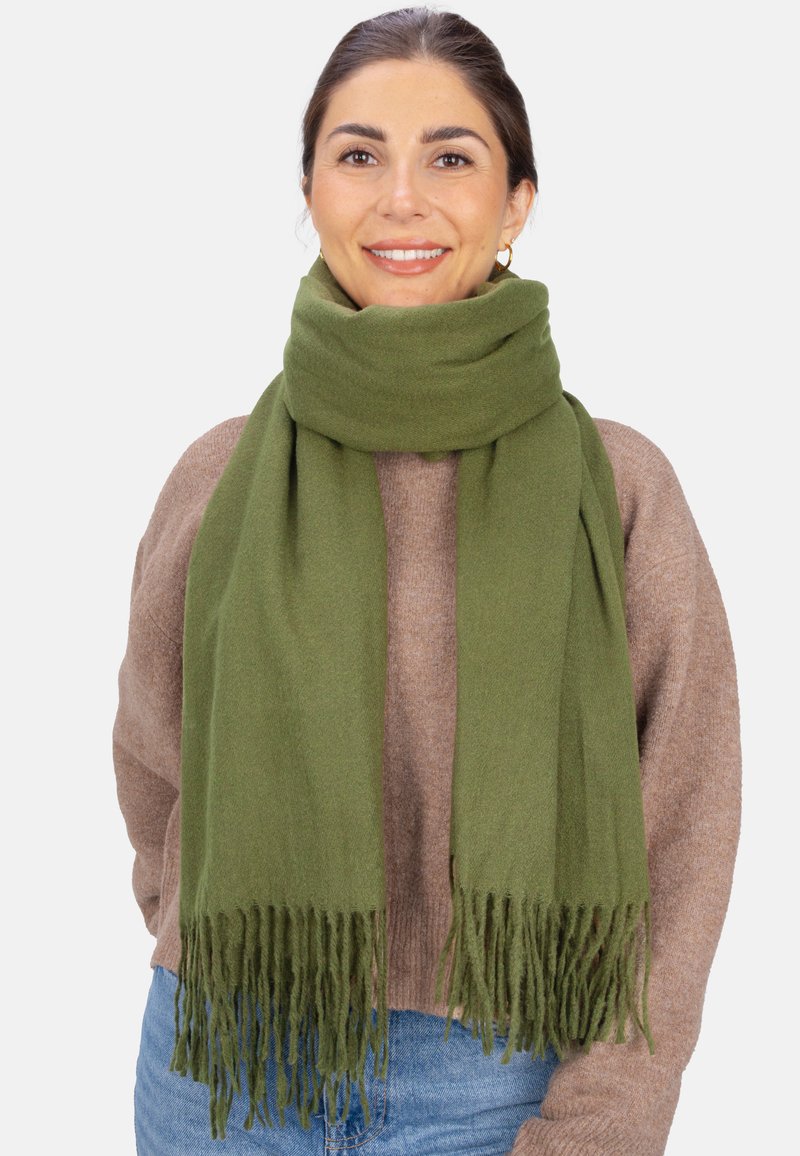 Écharpe vert olive en tissu doux, avec des bords frangés. Portée lâchement autour du cou sur un pull marron.