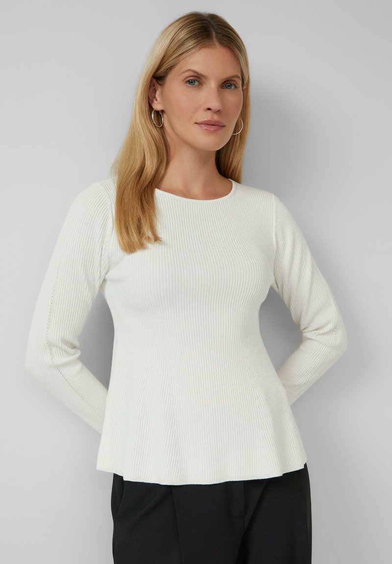 Haut long à manches longues blanc côtelé avec un col rond et un ourlet évasé, fabriqué à partir d'un tissu texturé. Un pantalon noir est partiellement visible en dessous.