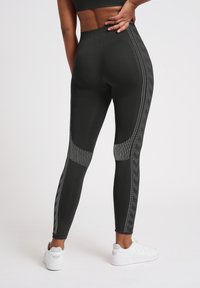 Person trägt schwarze Leggings mit hoher Taille und perforierten Seitenmustern sowie weiße Sneaker, Hand ruht im unteren Rückenbereich.