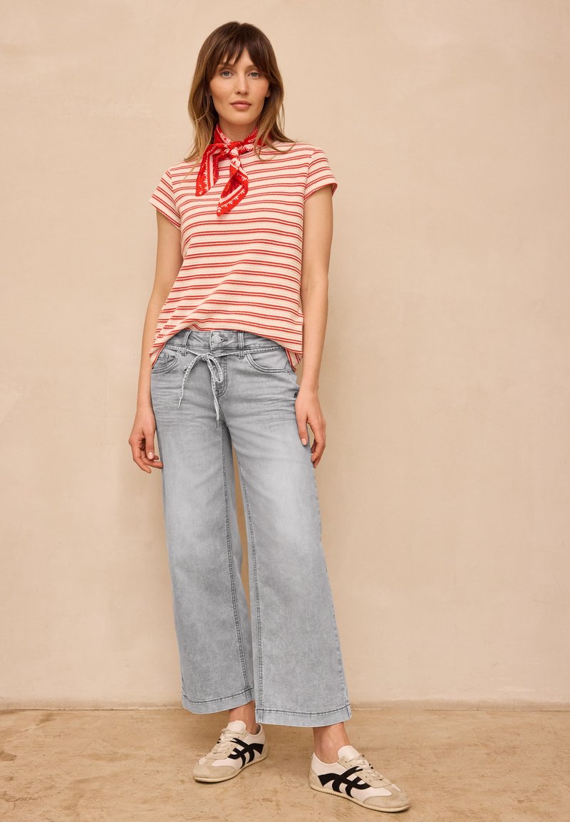 Femme portant un t-shirt à manches courtes rayé rouge et blanc, un foulard rouge autour du cou, un jean large gris clair et des baskets blanches avec des rayures noires.