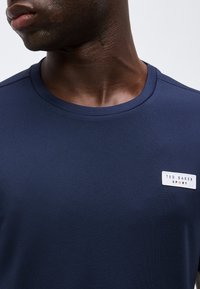 Κοντινό πλάνο ενός προσώπου που φοράει μπλούζα με στρογγυλή λαιμόκοψη σε ναυτικό μπλε χρώμα με μία λευκή ορθογώνια ετικέτα "Ted Baker Sport" στο στήθος.