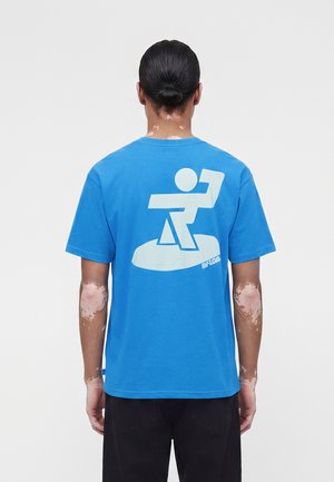 Vitiligós személy kék pólót visel, melyen hátul nagy fehér absztrakt grafika található, és fekete nadrágot, háttal áll.