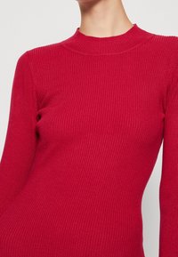Pull en maille côtelée rouge avec un col montant et des manches longues, présentant une coupe ajustée et une texture lisse.