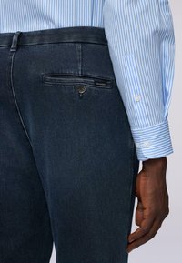 Pantalones de mezclilla oscura con un bolsillo trasero y un botón, combinados con una camisa a rayas azul claro y blanca con puños abotonados.