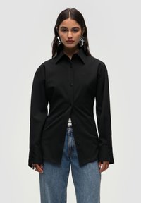Sort langærmet skjorte med knaplukning foran, der har en klassisk krave og subtil logo-brodering, parret med lyseblå jeans.