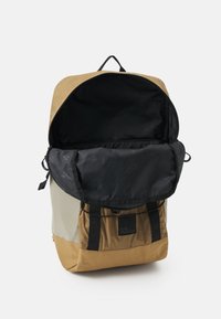 Rucksack mit einem beige und grauen Äußeren, einer schwarzen Oberklappe, verstellbaren Trägern und einem Logopatch. Das Innere besteht aus schwarzem Nylon.