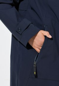 La veste imperméable marine est dotée d'une poche zippée, de manches à boutons et d'une texture lisse. La tirette de la fermeture éclair a un accent jaune pour une prise en main facile.