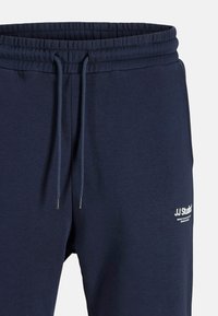Navy blue joggers with elastic waistband and adjustable drawstring. Features white branding on the left thigh. Smooth fabric texture.