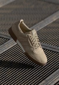 Sapatilha de camurça bege com bico arredondado, parte superior texturizada, atacadores combinando e sola de borracha castanha. Três riscas laterais em um tom mais claro.