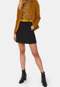 Černá minisukně, hořčicový blejzr, vzorovaný top. Blejzr má strukturovaný design, top má malé vícebarevné tečky, kotníčkové boty se zipem.