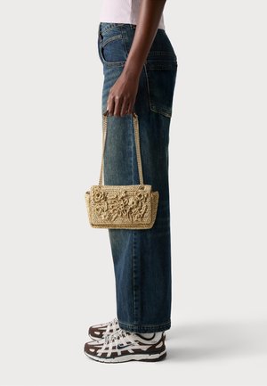 Eine Person in blauen Jeans und weißen Sneakers hält eine beigen gehäkelten Handtasche mit goldenen Kettenriemen an ihrer Seite.