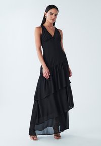 Juoda be rankovių maxi suknelė su gilia iškirpte ir sluoksniuota raukinio detalė, su pūstą, permatomą apatinę dalį.