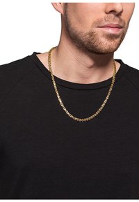 Gul metal kædema bracelet med sammenkædede led, båret over en sort struktureret skjorte, med en medium tykkelse og poleret finish.