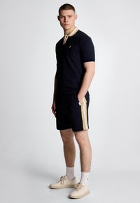Polo navy a maniche corte con colletto beige, dotato di un piccolo logo. Abbinato a pantaloni corti navy, evidenziati da una striscia dorata laterale e scarpe chiare.